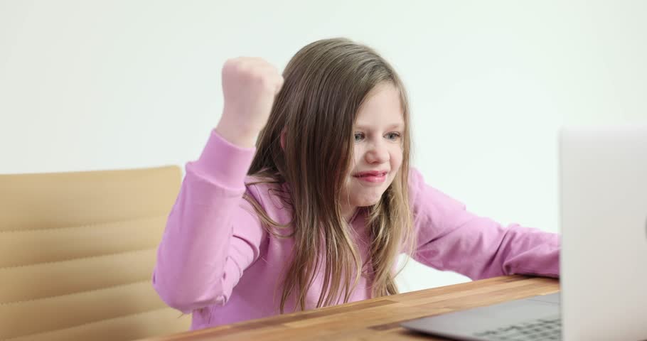 Preteen girl sits at table and stares intensely at laptop screen. Blonde child clenches fists tightly and pounds wooden desk in anger. Slow motion