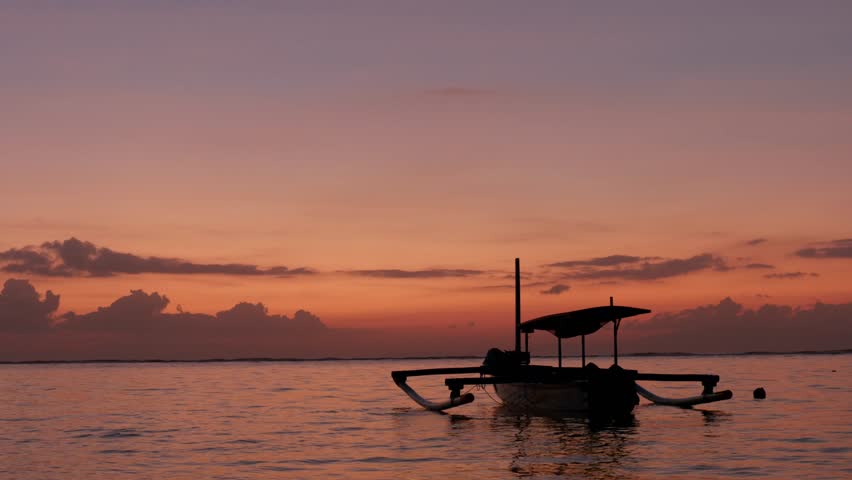 Sunrise on Sanur Beach, Bali. A traditional fishing boat sits at anchor. The local boat is silhouetted against the coloured sky. Clip 1