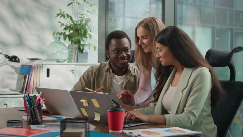 Happy African American casual informal businessman Arabian Indian businesswoman working talking using laptop teamwork Caucasian business woman come in office discuss online data multiracial colleagues