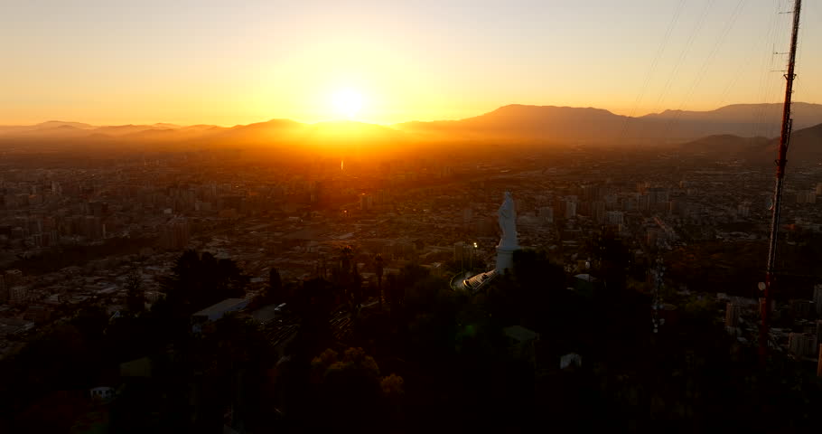 Drone dolly across Santiago from Cerro San Cristobal Christ statue over city with golden sky at dusk in Chile