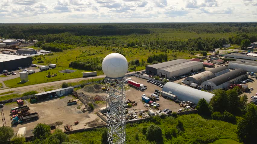 Aerial drone footage orbiting and flying around a modern weather observatory radar dome or white colored radome during a sunny summer day with landscape and city in the horizon at the background.