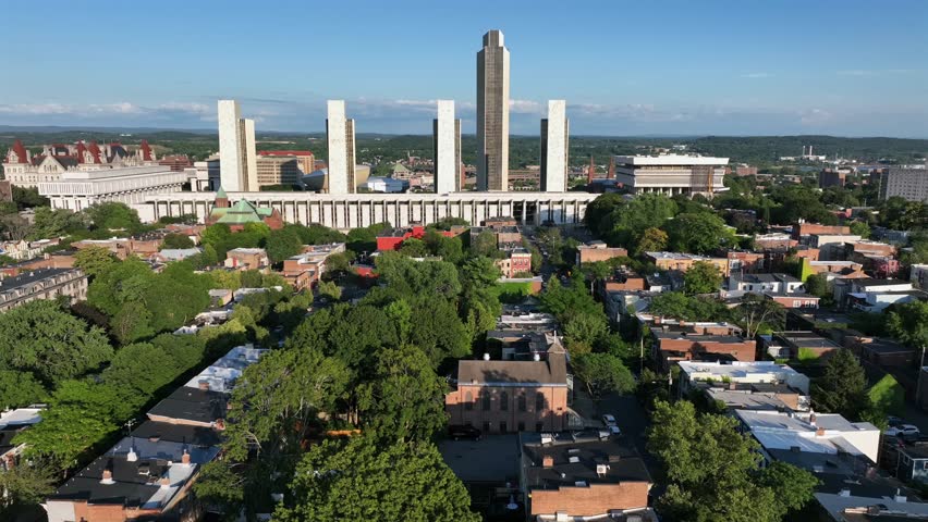 Idyllic green trees between brick houses and homes near Empire State plaza in New York. Aerial wide shot. Group of modern state office buildings in background. Peaceful neighborhood scene in summer.