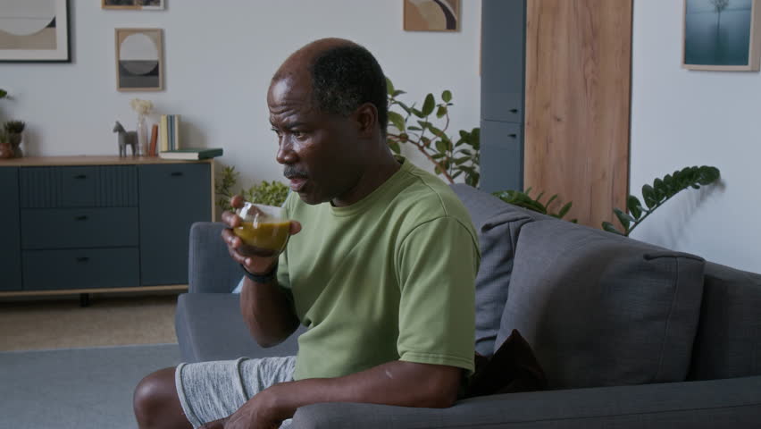 Medium shot of elderly African American man sitting on couch at home while resting after fitness workout, tasting and drinking glass of healthy freshly squeezed green vegetable or wheatgrass juice
