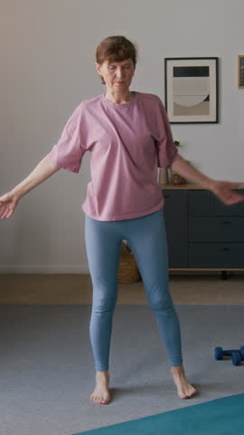 Full vertical shot of senior Caucasian woman in pink t-shirt and blue leggings stretching after fitness workout at home, bending forward, twisting at waist, during cool down