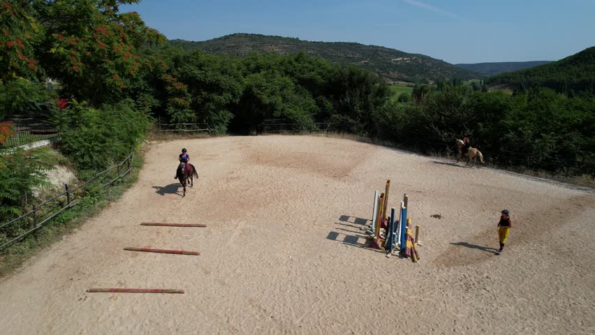 Pullback aerial view shows a trainer observing as a rider practices jumping between three ground bars