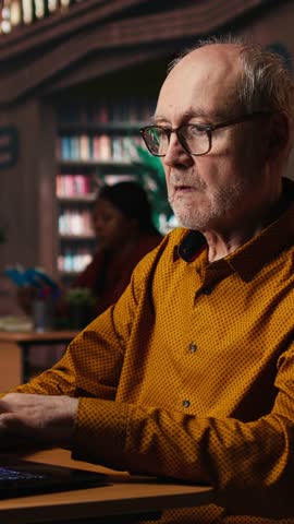 Vertical Video Focused senior guy reading academic text on laptop at the university, sitting in vintage study room and browsing online documents. Lifelong education through formal learning. Camera A.