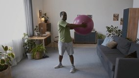 Wide shot of mature fit African American man standing in living room at home, doing torso rotation exercise while holding large fitness ball to maintain strength and mobility in old age - Powered by Shutterstock - Get 15% off with code: PIKWIZARD15