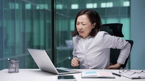 Tired asian businesswoman suffering from back pain while working on laptop sitting at workplace in business office. Upset female employee holds her hand on her lower back, massaging her sore muscles - Powered by Shutterstock - Get 15% off with code: PIKWIZARD15