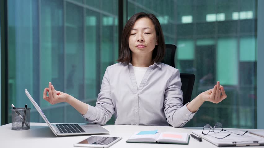Asian businesswoman meditates with her eyes closed sitting at workplace in modern business office. Female employee took a break at work. Calm woman rests, relaxes, relieves stress, feels peace of mind