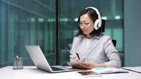 Asian businesswoman in headphones watching video call online conference taking notes using laptop sitting in office. Female employee listening remote business training talking communicates remotely - Powered by Shutterstock - Get 15% off with code: PIKWIZARD15