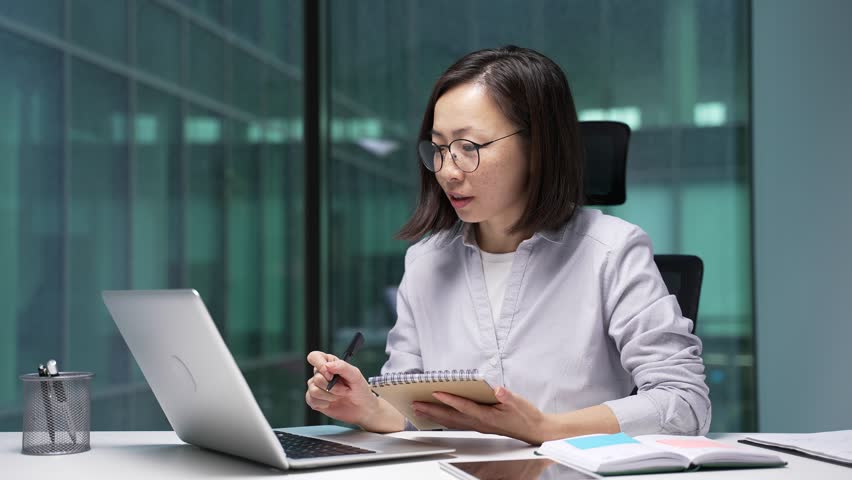 Asian businesswoman watching video call online conference taking notes in notebook looking at laptop screen sitting at workplace in office. Focused female employee listening remote business training