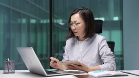 Asian businesswoman watching video call online conference taking notes in notebook looking at laptop screen sitting at workplace in office. Focused female employee listening remote business training - Powered by Shutterstock - Get 15% off with code: PIKWIZARD15