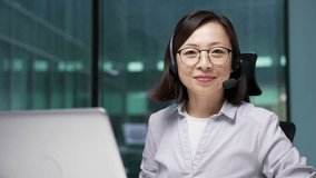Portrait of a happy Asian female corporate operator in a headset sitting at a workplace working on a computer in business office. Smiling female agent of call center posing looking at camera. Close up - Powered by Shutterstock - Get 15% off with code: PIKWIZARD15