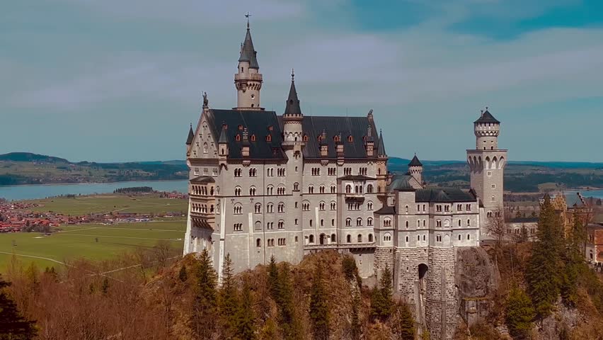 Historic Neuschwanstein Castle in Schwangau German on hilltop n mountain peak over grassland n road street with cars n river or lake on blue sky, 4k aerial view of famous landmark n nature landscape
