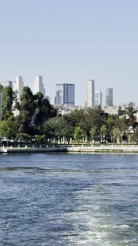 Static skyline of Istanbul downtown with modern skyscrapers and green trees. View from yacht floating away on calm Bosphorus. Glimpse of Yıldız Palace’s historic facade. Cinematic reel-style cityscape