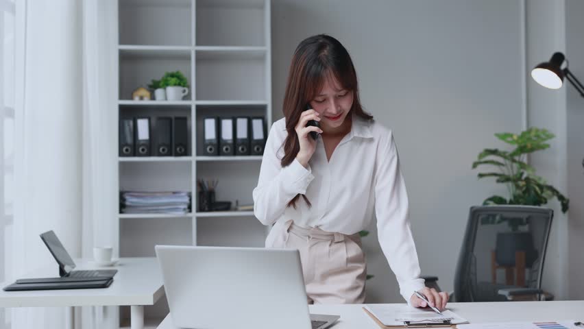 Young Asian businesswoman standing at her desk in a modern office, talking on her smartphone while reviewing documents and working on her laptop