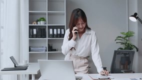 Young Asian businesswoman standing at her desk in a modern office, talking on her smartphone while reviewing documents and working on her laptop - Powered by Shutterstock - Get 15% off with code: PIKWIZARD15