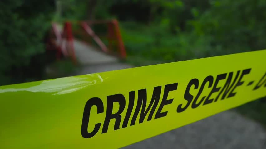 Close up view of yellow police line Crime Scene forensics investigation tape pulled in front of a nature rural road area leading to a small red bridge going over a river that is bokeh blurry. Daytime.