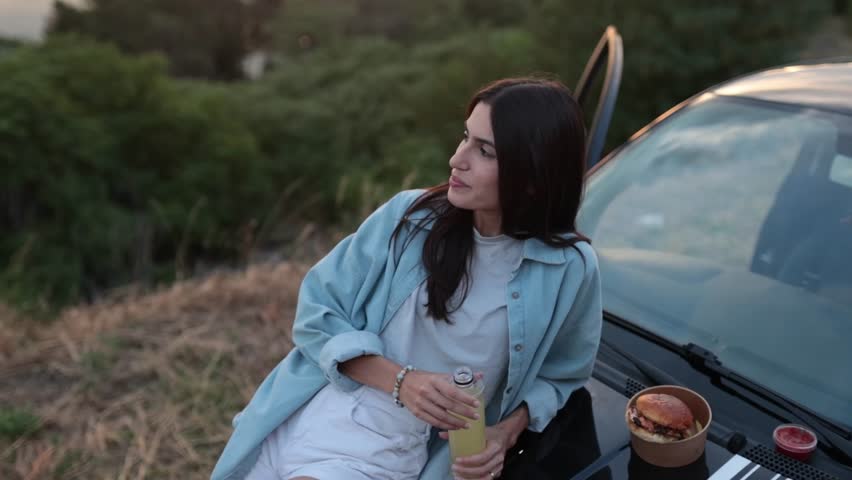 Happy woman enjoying a picnic near the river, eating french fries, drinking lemonade and having a burger during sunset. 4k video