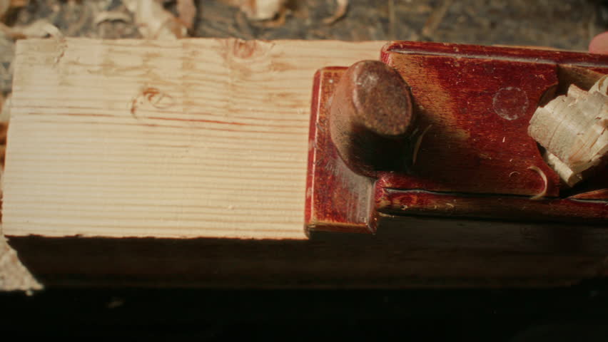 Top down close up view of hand of woodworker planing wood with traditional handheld planer, producing curled shavings while smoothing timber surface