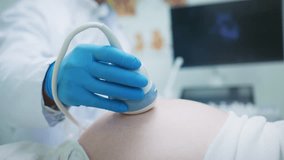 Close up gloved hand of doctor holding ultrasound probe. Scanning belly of pregnant woman. Medical examination taking place in hospital room. Monitor with image in background. Evaluation. - Powered by Shutterstock - Get 15% off with code: PIKWIZARD15