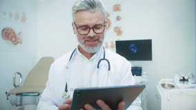 Close up portrait Caucasian male doctor with gray hair and beard wearing glasses and stethoscope reading on tablet. Medical specialist analyzing patient information in hospital office. - Powered by Shutterstock - Get 15% off with code: PIKWIZARD15
