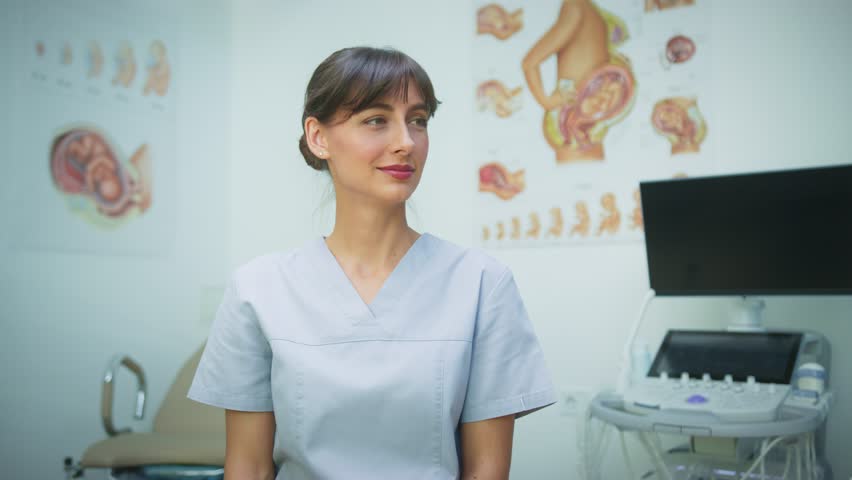 Caucasian female doctor with dark hair wearing medical scrubs. Smiling while looking at camera in hospital office. Healthcare professional sitting near ultrasound. Ready for patient consultation.