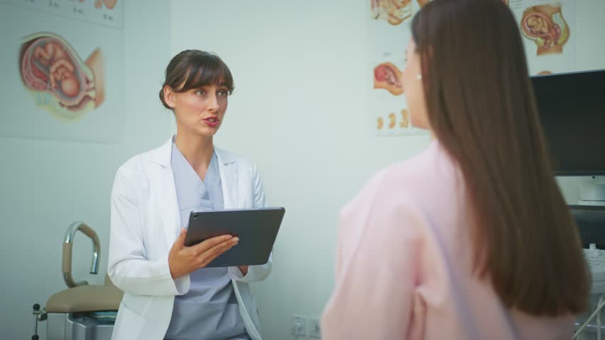 Caucasian female doctor in white coat holding digital tablet smiling while talking to patient. Medical specialist giving positive news after check up in gynecology office. Hospital.