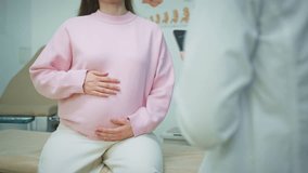 Beautiful pregnant Caucasian woman in pink sweater rubbing belly while talking to doctor. Expectant mother discussing health during prenatal consultation in gynecology office. Indoors. - Powered by Shutterstock - Get 15% off with code: PIKWIZARD15