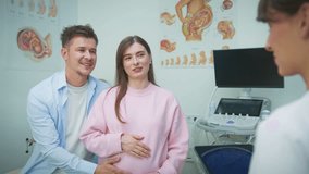 Lovely Caucasian couple expecting baby sitting together in gynecology office. Woman in pink sweater holding belly while man embracing her. Talking with doctor during consultation. Indoors. - Powered by Shutterstock - Get 15% off with code: PIKWIZARD15