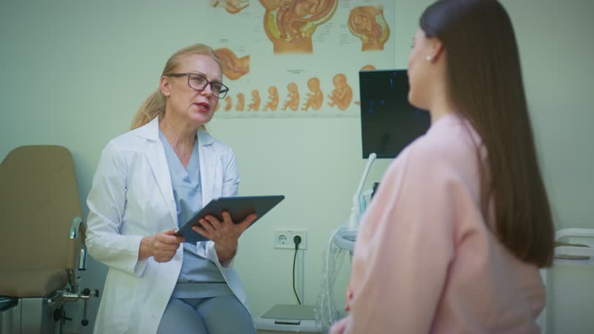 Caucasian doctor in white coat writing notes on digital tablet while talking with pregnant patient. Medical professional documenting examination results in consultation room with posters.