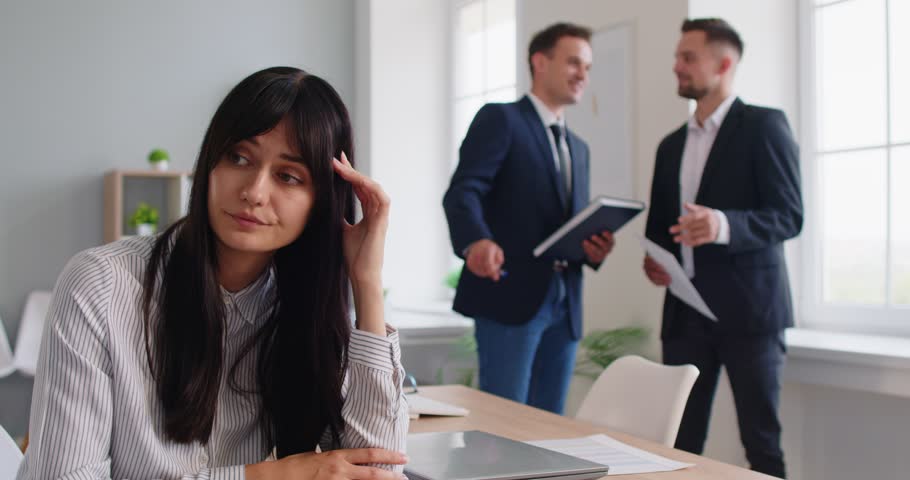 A businesswoman rubs her head at a desk as colleagues talk showing stress, anxiety, and isolation from overwork, conflict, deadlines, and fatigue. Pensive employee with headache in bright office.