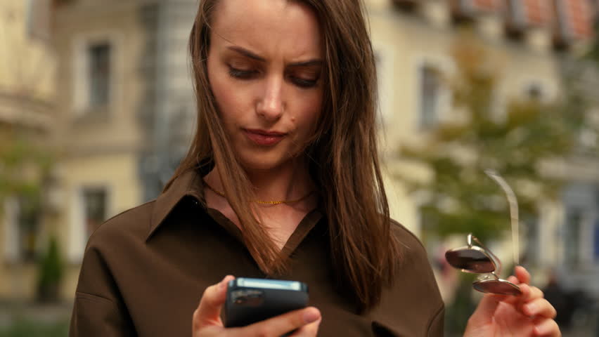 Confused Caucasian woman with mobile phone lost direction in city searching location address outdoors using navigator looking around female using smartphone GPS service girl at street hold sunglasses