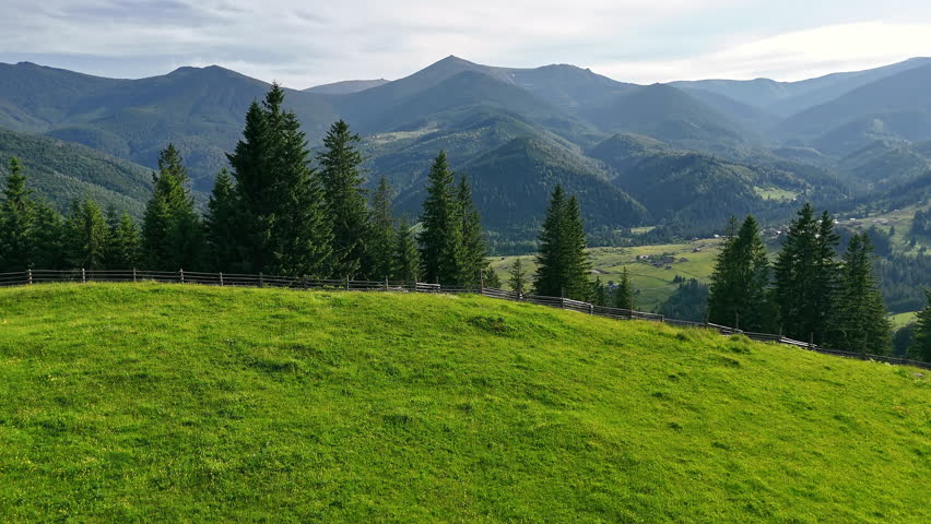 Mountain landscape with pine trees and village valley. Lush green hillside with tall pine trees overlooking a scenic valley dotted with houses and surrounded by rolling mountain peaks.
