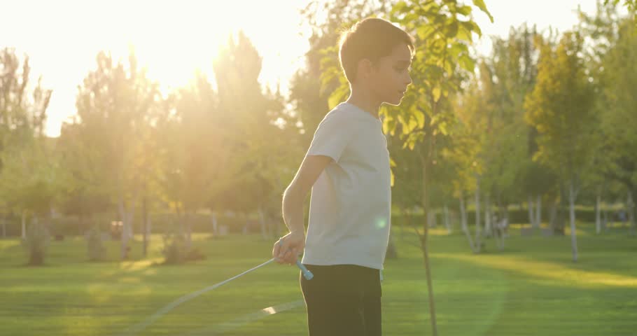 Energetic happy kid jumps rope in sunny park during golden hour. Kid enjoys outdoor exercise movement freedom in nature sunset. Joyful kid active play wellness. Summer fitness healthy lifestyle child.