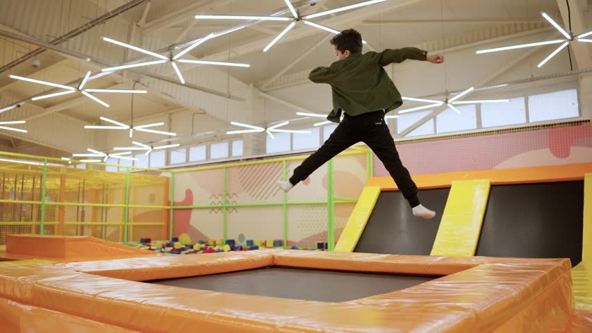 Joyful happy African-American boy jumping on trampoline in colorful kids play center, having fun. Slow motion