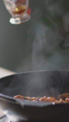 Vertical close-up shot of unrecognizable male hands precisely pouring alcohol into skillet with chicken pieces before carefully igniting it with lighter