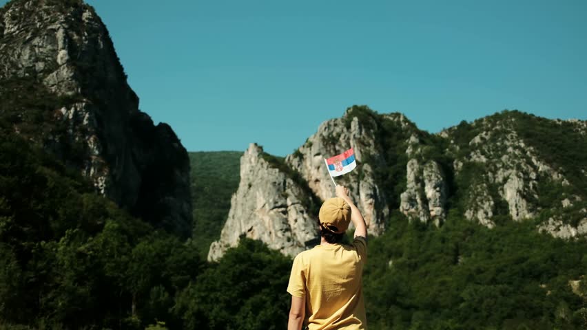 Female traveler in a yellow shirt waves the Serbian flag towards dramatic mountain peaks, celebrating culture and adventure in nature. Rear view. Stara Planina national park, Serbia country