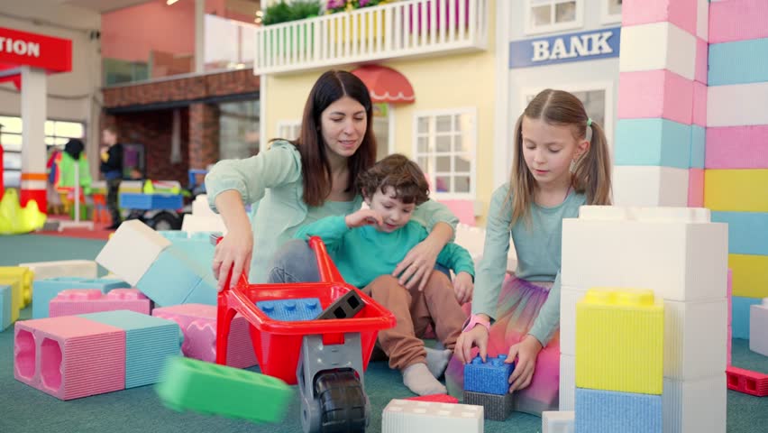 Smiling pretty young mother and her kids playing together in the game center while building with big foam plastic blocks. Slow motion