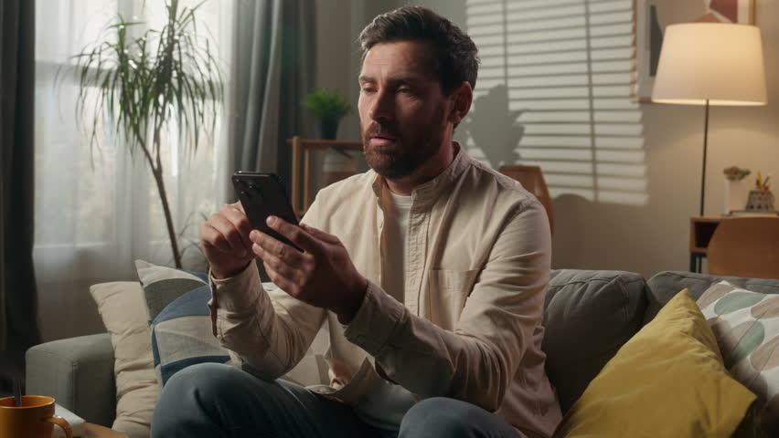 Sad upset frustrated man Caucasian businessman guy bored using mobile phone on couch sofa worried disappointed male homeowner reading news cellphone browsing message scam scrolling smartphone at home