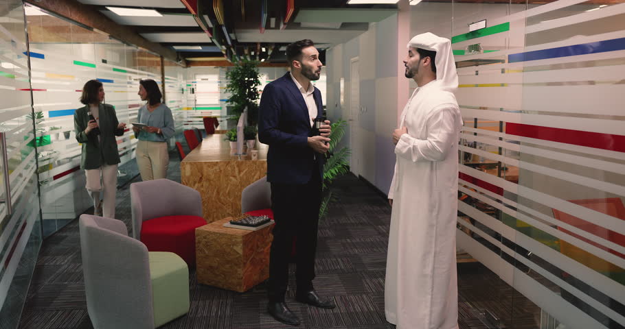 In cozy lounge area of modern co-working office multinational team members lead casual conversation during workday break. Arabic man dressed in white thobe talks with Hispanic colleague. Communication