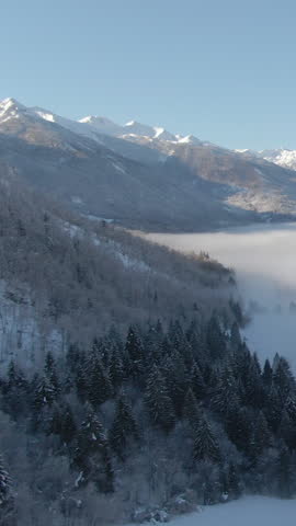 DRONE: Dense white fog covers the valley under the spectacular snowy mountain range in the Julian Alps. Breathtaking aerial view of towering mountaintops and forests near picturesque Bohinj, Slovenia.