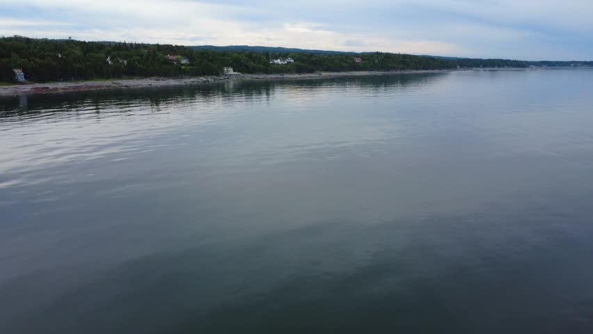 Panoramic aerial video of a peaceful coastline with a beach, houses, wooded areas, and calm waters under a cloud-darkened sky. St. Lawrence River, Quebec, Canada.