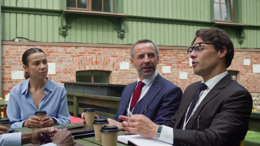 Five confident intercultural delegates in formalwear sitting by table in outdoor cafe at break and looking at young African American colleague explaining details of his speech at conference