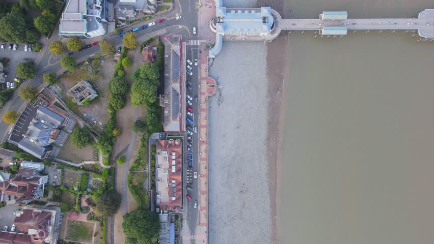 Aerial top down shot of a road following the coastline moves over Penarth Pier before stopping over a Jetski docking on the beach, lots of people and cars below, waves on water, Wales, UK, 4K, 60fps
