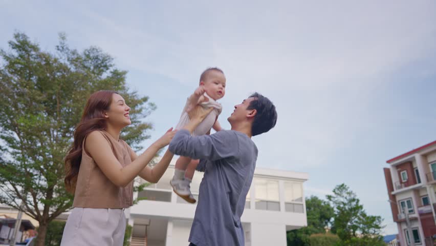 Asian young parents holding and gently tosses baby boy outdoors in park. Attractive couple share a tender moment playing with newborn child, fostering a warm and loving family atmosphere in the garden