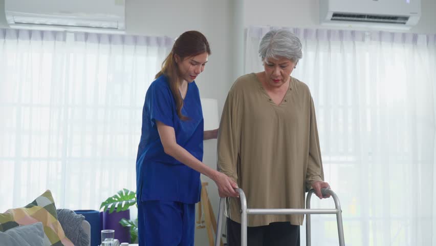 Asian senior older woman patient doing physical therapy with caregiver. Attractive specialist carer help and support elderly mature grandma practice walking slowly with walker at nursing home care.