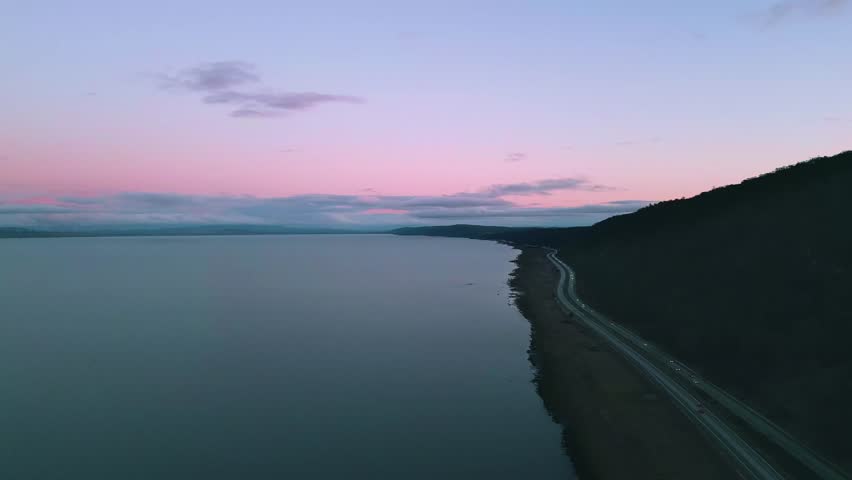 Pink night sky over the lake with highway and cars with headlights on passing below. Wind turbines line the lefthand side of the lake. 