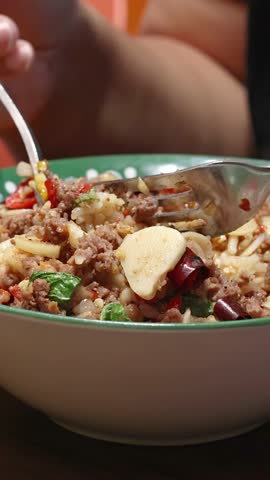 Close-up of a person using a spoon and fork to eat a colorful Thai beef, garlic, chili, and rice meal in a decorative bowl under warm lighting