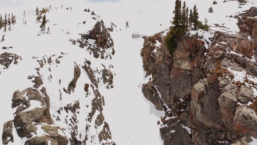 Snowboarder turning down cornice rock walls chute Deer Creek Trail Montezuma winter Rocky Mountain Colorado avalanche terrain aerial drone sunny blue sky deep powder snow snowboard backcountry follow